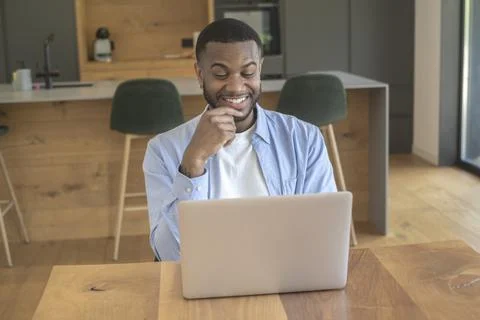 Young man smiling at laptop while working from home in a modern kitchen, en.. Stock Photos
