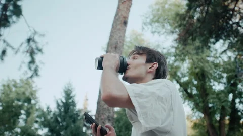 Young man smiling while drinking coffee and using phone outdoors in park with Stock Footage 340859078