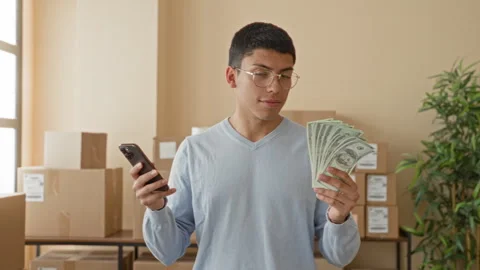 Young man smiling while holding fanned dollar bills and a smartphone beside.. 스톡 동영상 337431591