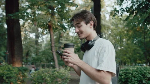 Young man smiling while walking in park, sipping coffee and using phone with Stock Footage 340859071