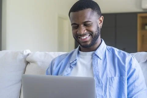 Young man smiling while working on a laptop at home, relaxed on a couch and.. Stock Photos