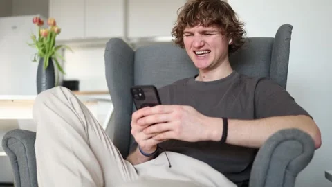 A young man with a smirk on his face sits in a chair, holding a phone and lookin Stock Footage 302633712