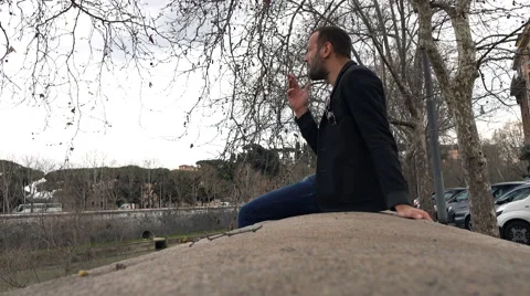 Young man smoking cigarette while sitting on ledge in city  Stock Footage 67089391