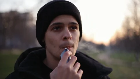Young man smoking/vaping with help of device for heating tobacco in the park in Stock Footage 121061941