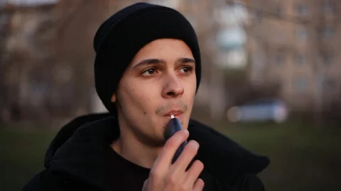 Young man smoking/vaping with help of device for heating tobacco in the park in Stock Footage 121061995