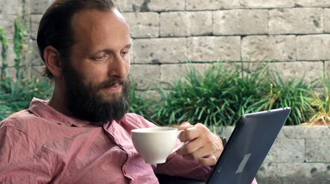 Young man on sofa reading article on tablet and drinking coffee on patio Stock Footage 63022370