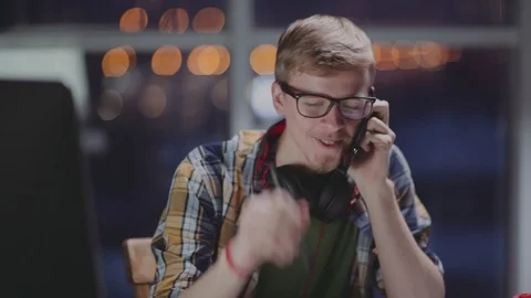 Young man speaks on the phone at the computer in front of a window. Stock Footage 87160902