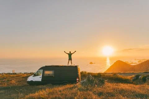 Young man on sprinter van with arms wide open in front of sunset on cliff in Stock Photos