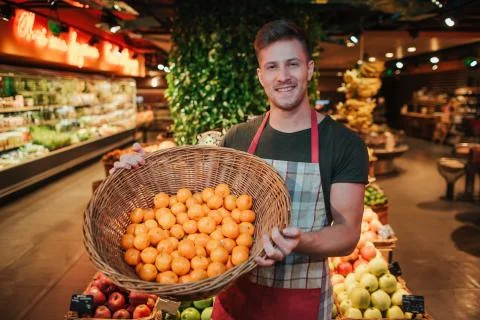 Young man stand at fruit boxes in grocery store. He hold basket with oranges and Foto stock