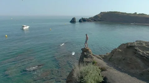 Young man is standing on a cliff above a clear sea with boats on the background Stock Footage 127280158