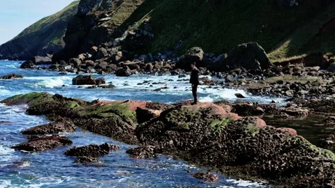 Young man standing on mossy rocks above ocean tide. Teenager watching waves near Stock Footage 320399377