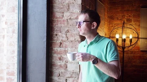 Young man standing near a window in a cafe and drink coffee or tea, breakfast in Video stock 73127265