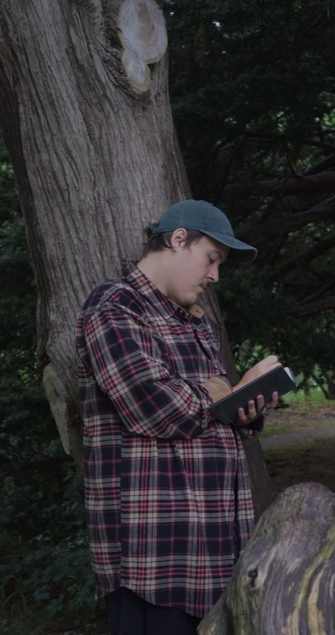 Young man standing peacefully under a large tree, journaling in a notebook. Vidéo 313949908