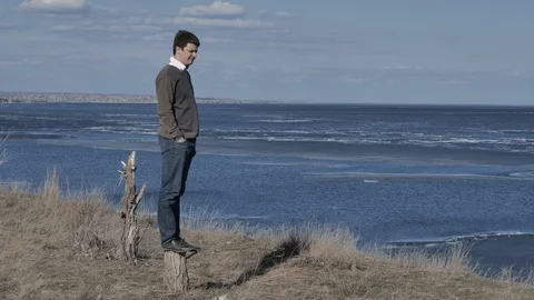 A young man is standing on a stump and thinking,in background a river,sunny day Stock Footage 124749396