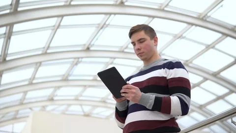 A young man is standing with a tablet. A man uses a tablet against a pan-glass Video stock 127393844