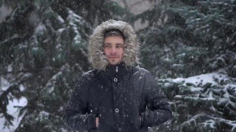 A young man stands against a background of fir trees under heavy snowfall in Stock Footage 257198747