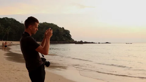 Young man stands on the beach by breaking waves and shoots video on a smartphone Stock Footage 146012884