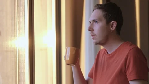 A young man stands next to a window sips from a coffee mug looking out window Stock Footage 276912781