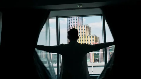 Young man stands by the panoramic window and opens the curtain Stock Footage 246583610
