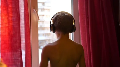 The young man stands by the window and dances, listening to music in headphones 库存影片 87504153