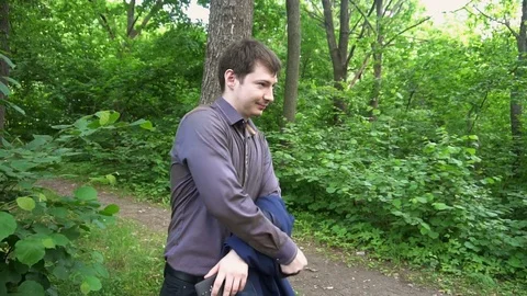 A young man stands in the woods,throws the jacket over his shoulder and makes a 스톡 동영상 77143068