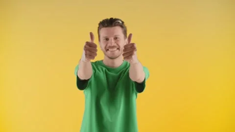 A young man stands on a yellow background and raises his hands showing thumbs Stock Footage 240318146