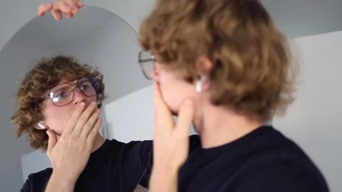 Young man stares into the mirror with a skeptical look as if questioning his own Stock Footage 312981927