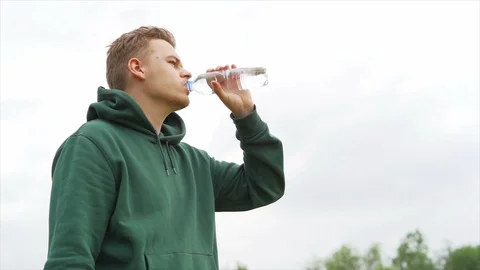 Young man is staring away from camera in hoodie and drink water from bottle Stock Footage 91230930