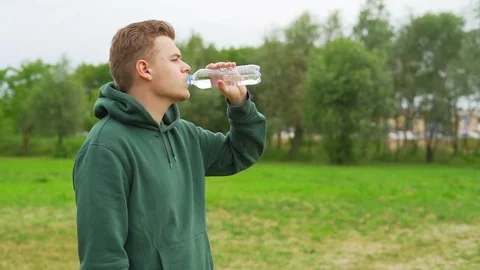 Young man is staring away from camera in hoodie and drink water from bottle Stock Footage 91230954