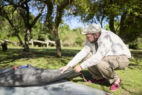 Young man starting the process of setting up his tent at a peaceful campsite. Stock Photos