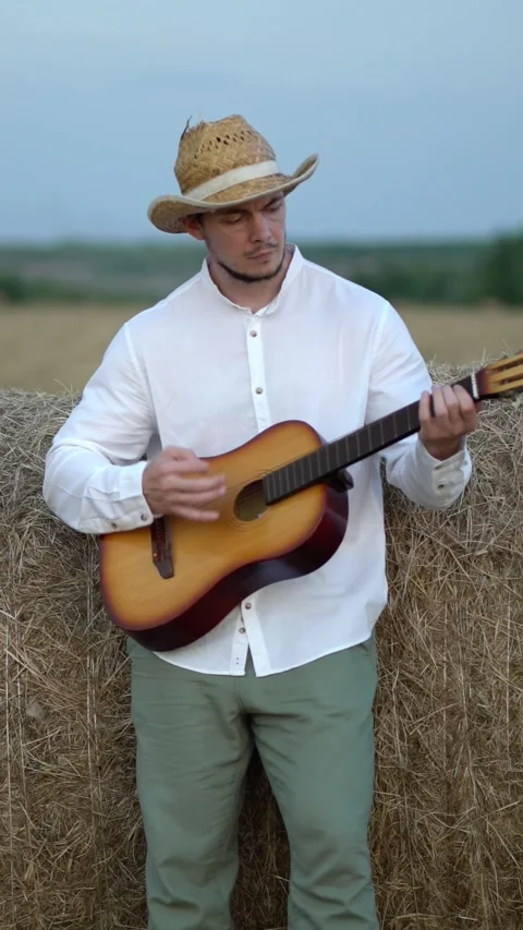 Young man in straw cowboy hat plays acoustic guitar leaning on hay bale in rural Stock Footage 341530321