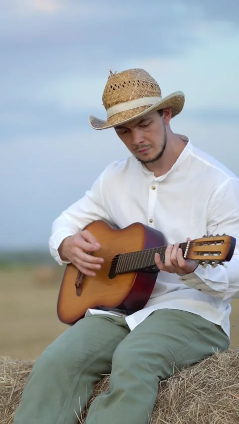 Young man in a straw hat plays acoustic guitar while sitting on a hay bale Video stock 342570623