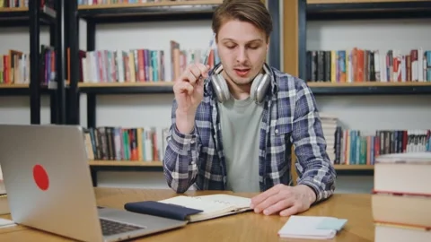 Young man studying in library with laptop, headphones, and notebook during study Stock Footage 318089758