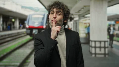 Young man in stylish attire making hush gesture at busy train station with .. Stock Footage 307324150