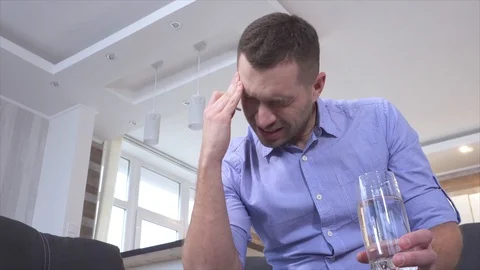 Young man suffer from hangover or headache. Sit alone and put white tablet into Video stock 128911655