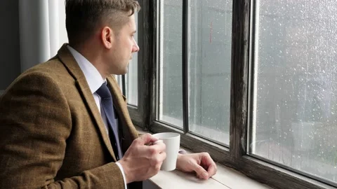 A young man in a suit drinks tea while standing by the window during the rain. Stock Footage 132180726