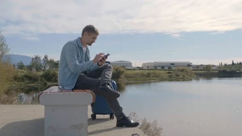A young man with a suitcase sits on a bench near the lake and uses the phone. Stock Footage 119324192