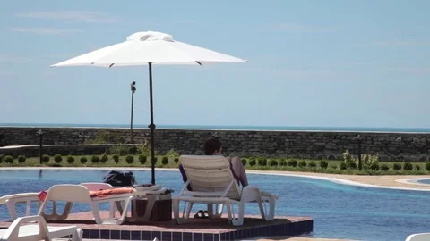 Young man on a sunbed with umbrella by the pool looking in the distance Stock Footage 69784517