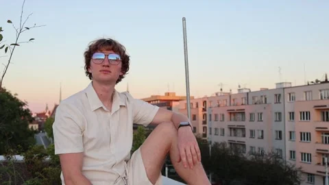 Young man in sunglasses sitting on rooftop at sunset calm sky and warm light ove Stock Footage 315413848
