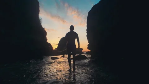 Young man with a surfboard, between the rocks by the ocean. Slow motion. Stock Footage 157991537
