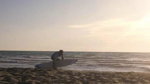 Young man surfer in wetsuit picks up surf board at the beach walking on the sand Stock Footage 85388596
