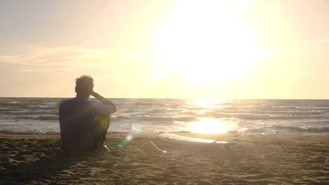 Young man surfer in wetsuit sitting with surf board lying down on the sand at Stock Footage 85452982