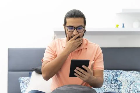 Young man surprised while reading sitting on bed Stock Photos