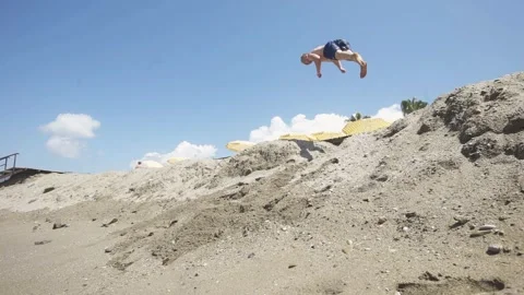 Young man in swim trunks leaps from sandy hill towards the beach, showcasing Видео 334672702