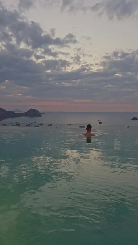 Young man swimming in infinity pool overlooking ocean at sunset Stock Footage 347141139