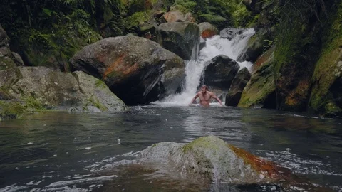 Young man swimming in river stream flowing from waterfall in tropical forest Stock Footage 107637322