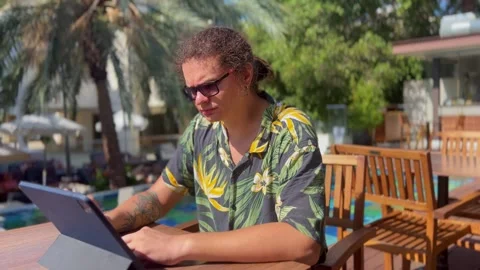 A young man swipes on a tablet screen while sitting by the pool in beachwear Video stock 288579518
