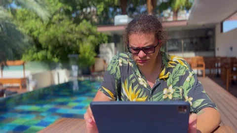 A young man swipes on a tablet screen while sitting by the pool in beachwear Stock Footage 288579536