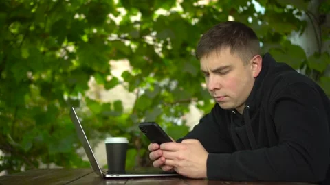 A young man at a table in a gazebo is texting on a smartphone during the day. A Stock Footage 251726750