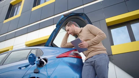 Young man with tablet device waiting till his electric car charge Video stock 247454497